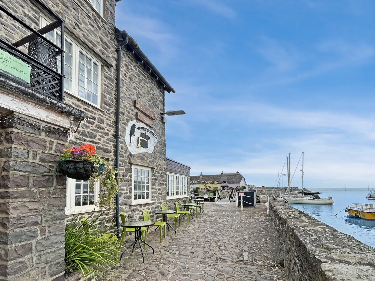 Harbour House, a stone building on the cobbled quay at Porlock Weir, with an iron balcony on its upper floor; café tables along the quay and boats in the harbour beyond