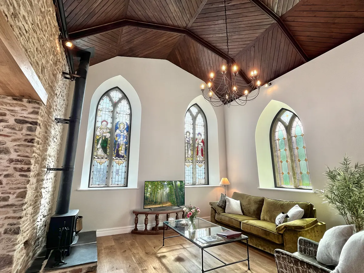 The nave of a converted chapel, with arched stained-glass windows, a wood-burner and a green velvet sofa under a timber roof
